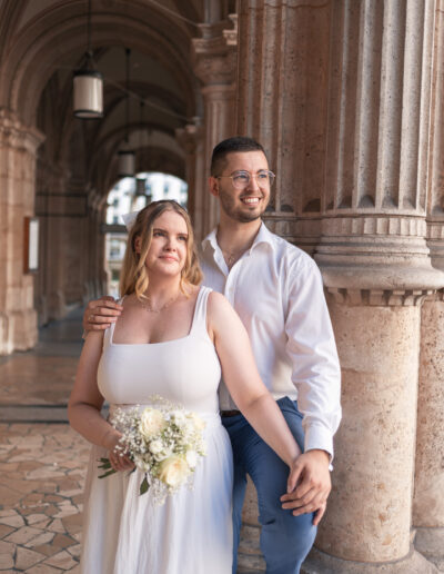 Hochzeitsfoto, vor der Wiener Staatsoper, Paar steht an eine Säule gelehnt und schaut verliebt in die Ferne, Braut trägt einen Brautstrauß in der Hand