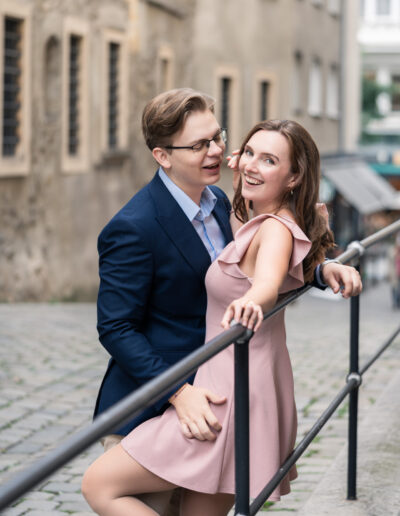 Paar steht am Gelände, Mann schaut Freundin an, Frau lächelt in die Kamer. Mann trägt Anzug, Frau alsros Kleid. Ort: Wien Innere Stadt