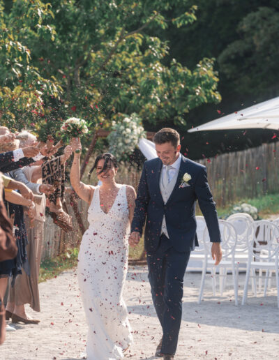 Hochzeitsfoto, Paar geht durch das Spalier und wird mit Blumen beworfen, Braut hält Brautstrauß nach oben, sie lächeln.