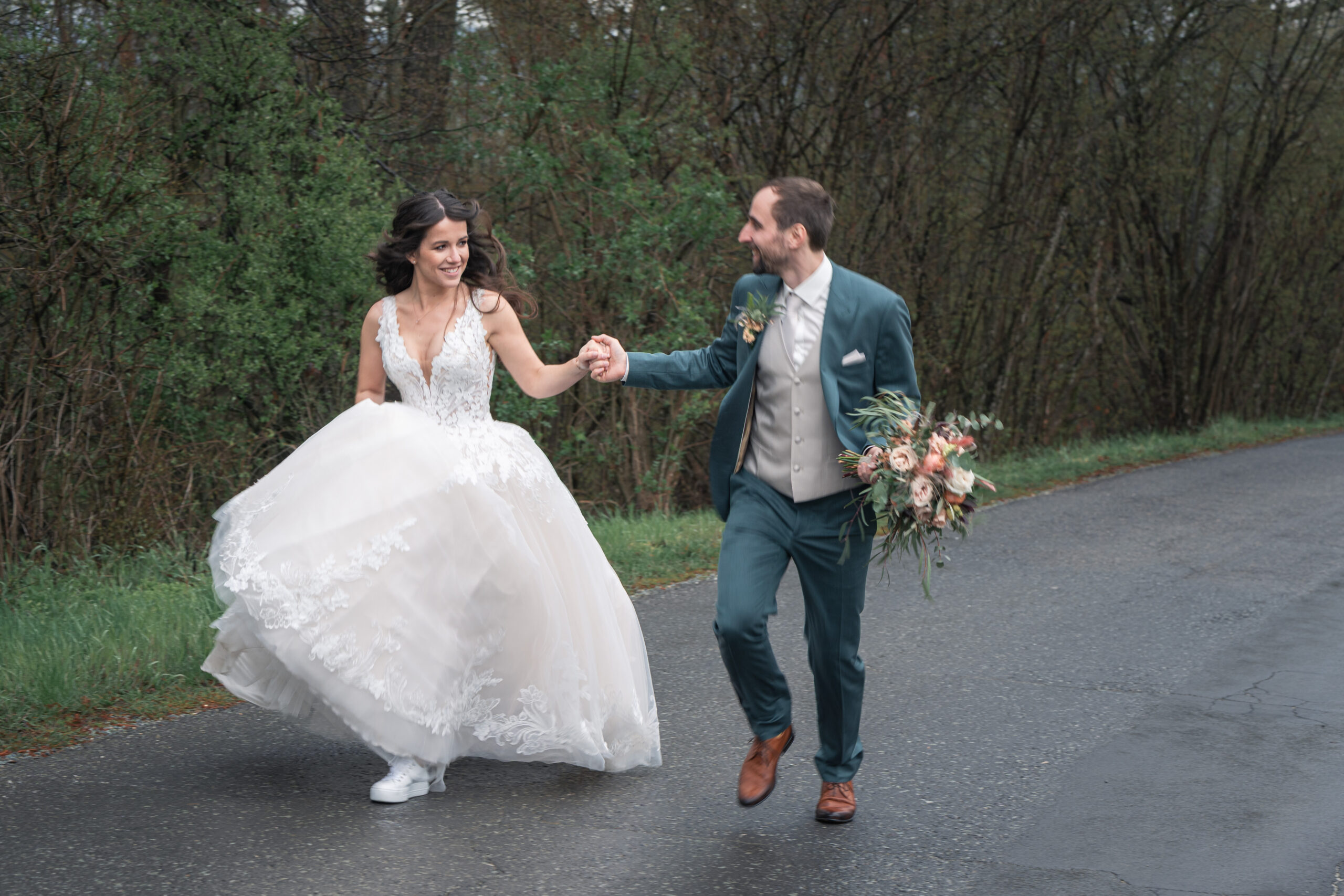 Hochzeitsfoto: Hochzeitspaar läuft die Straße entlang Braut trägt Brautgleit und hält Bräutigam an der Hand der ihren Brautstrauß trägt. Ort: Am Hannersberg